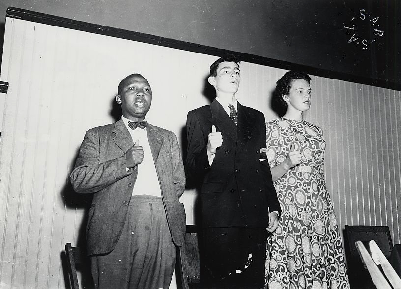 Albie Sachs with Joseph Nkatlo and Mary Butcher giving the thumbs-up sign while singing "Nkosi Sikilel' iAfrika" at a Defiance Campaign meeting (April 12, 1952). The fist signifies strength in unity and the upright thumb signifies optimism that the struggle will succeed. Sachs and Butcher were among the first white South Africans to join the campaign in Cape Town. (National Library of South Africa: Cape Town; Cape Times Negative Collection)